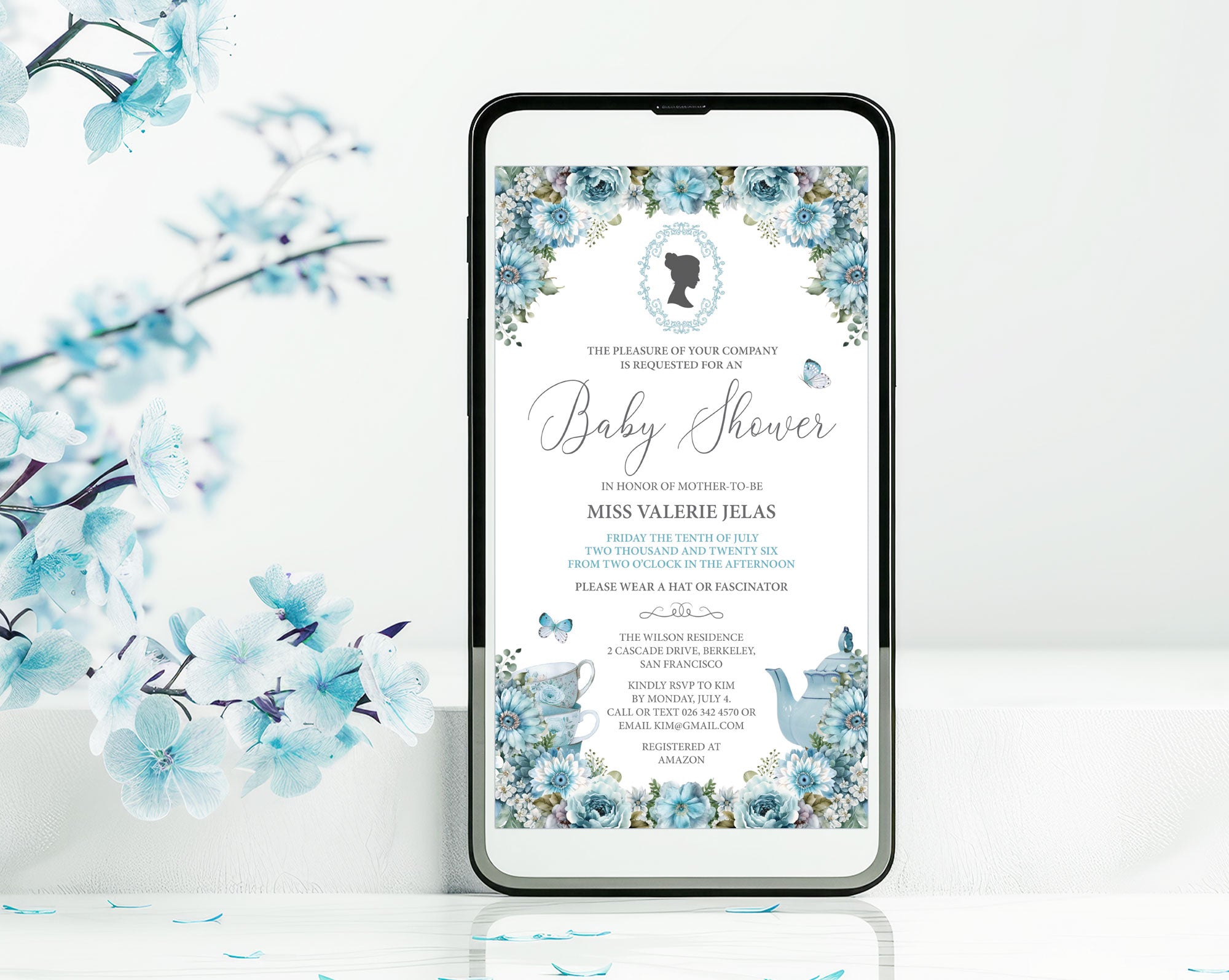 a black mobile phone with a dusty blue flower baby shower tea invitation showing on the screen, leaning up against a white wall with some blue blossoms