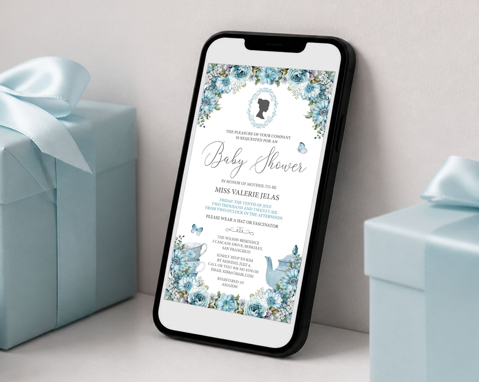 a black mobile phone showing a blue flower baby shower regency style invitation leaning up against a white wall next to some blue wrapped gifts
