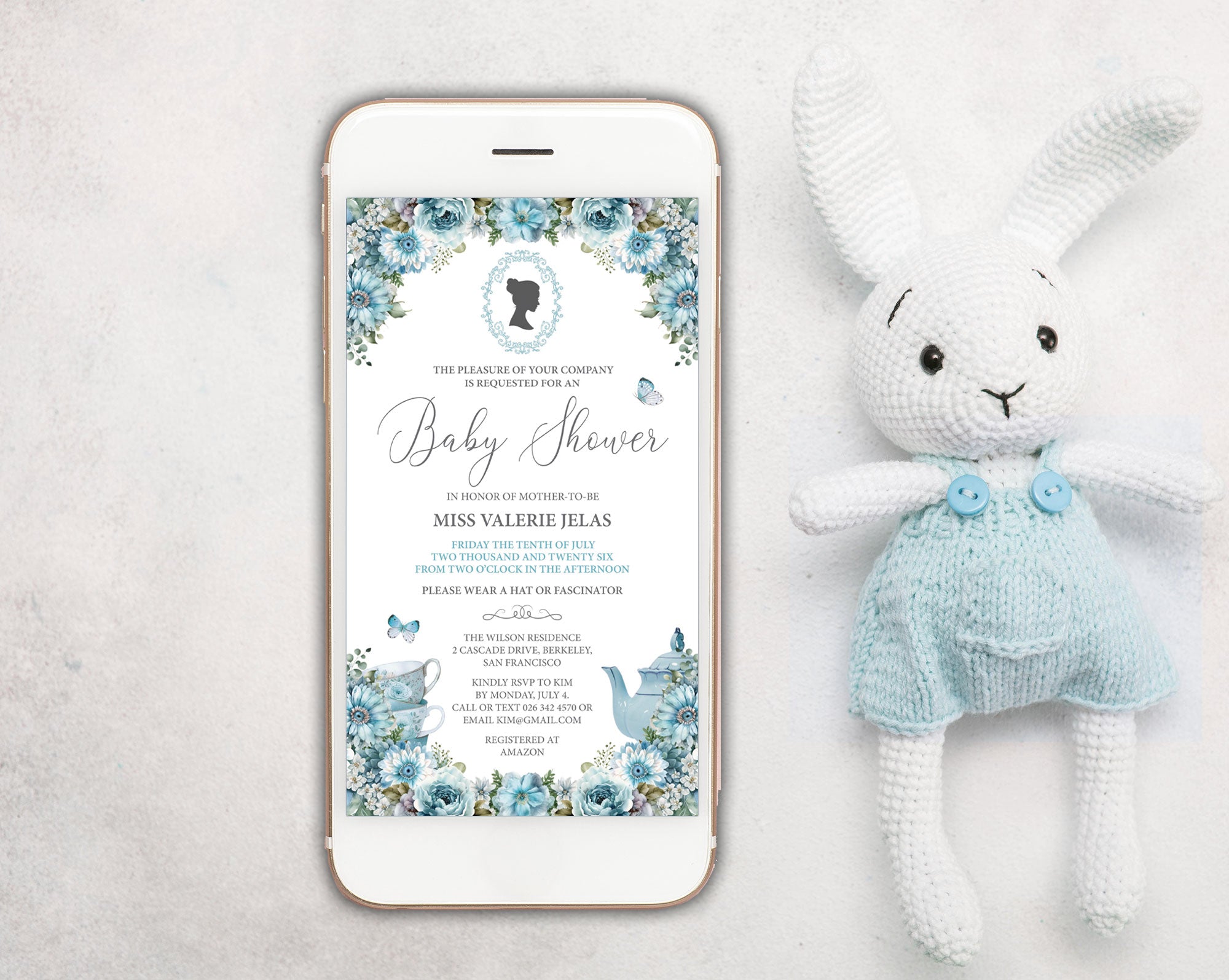 a dusty blue baby shower invitation showing on the screen of a white mobile phone, laying on a white background next to a stuffed rabbit with blue dungarees