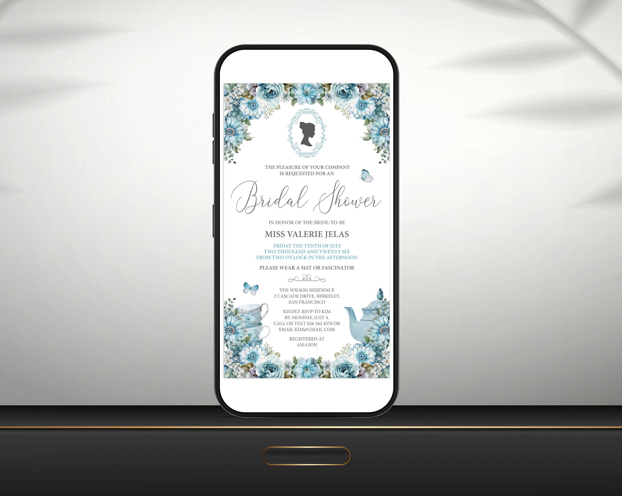 a black smart phone with a dusty blue regency style bridal shower tea invitation showing on the screen, leaning up against a white wall sitting on a black shelf