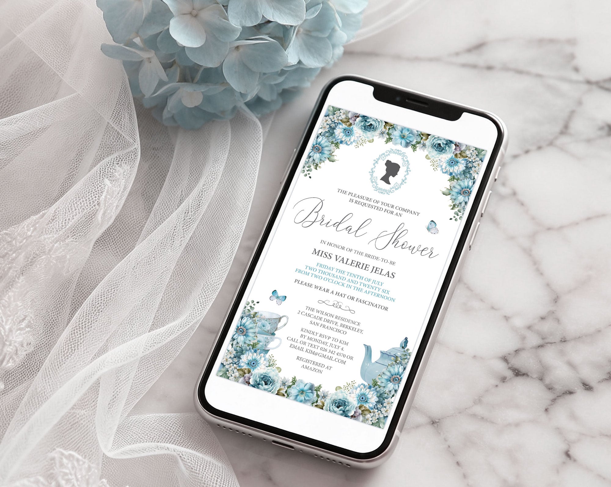 a black mobile phone with an elegant blue flower regency bridal shower invitation showing on the screen laying on a marble surface with a bridal veil and a blue hydrangea flower