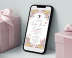 a black mobile phone showing a pastel flower baby shower regency style invitation leaning up against a white wall next to some pink wrapped gifts