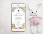a pastel baby shower invitation showing on the screen of a white mobile phone, laying on a white background next to a stuffed rabbit with pink dungarees