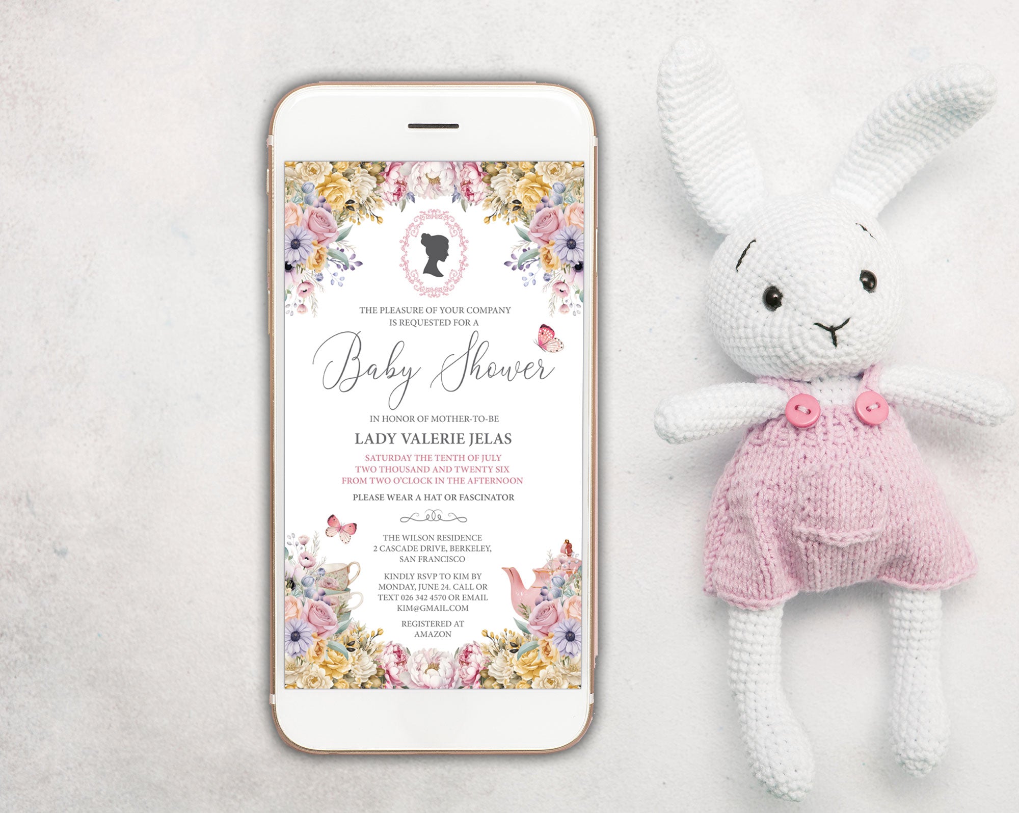 a pastel baby shower invitation showing on the screen of a white mobile phone, laying on a white background next to a stuffed rabbit with pink dungarees