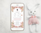 Phone displaying a blush pink baby birthday floral invitation with a white rabbit toy wearing a pink dress on a light gray background