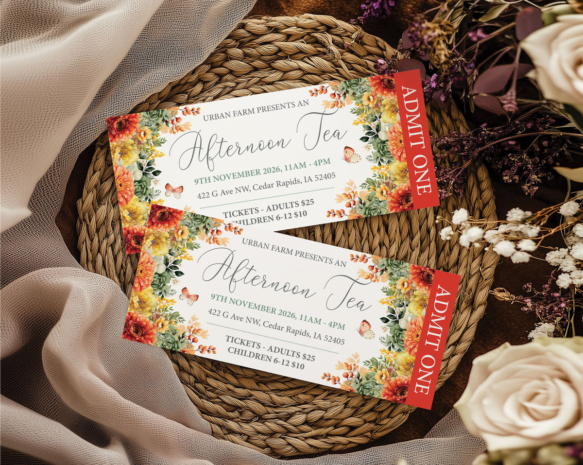 Two printed event tickets for an afternoon Tea event are arranged on a woven round tray with draped fabric and dried flowers around them. The tickets have floral borders in orange, yellow, green, and red, with red side panels reading ADMIT ONE.
