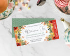 A printed tea invitation card with floral borders and event details, placed on top of a green patterned envelope on a white marble surface with tea and pastry props around it.