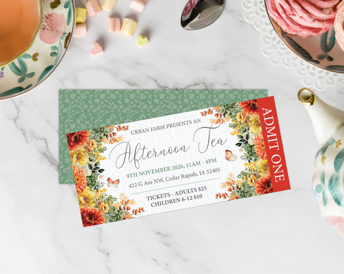 A printed tea invitation card with floral borders and event details, placed on top of a green patterned envelope on a white marble surface with tea and pastry props around it.
