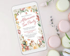 elegant floral afternoon tea invitation showing on a white mobile phone which is laying on a white background surrounded by macarons
