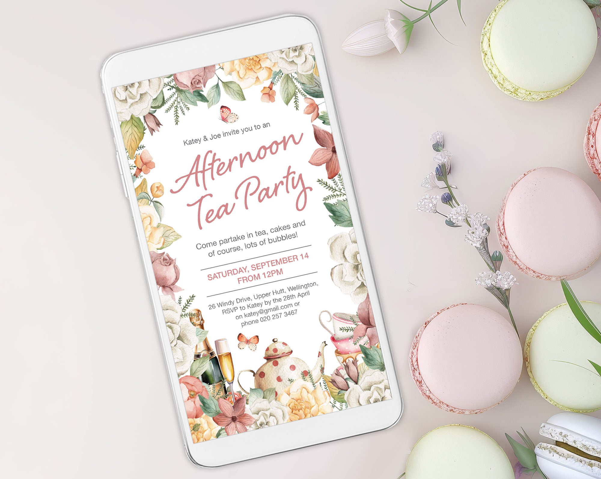 elegant floral afternoon tea invitation showing on a white mobile phone which is laying on a white background surrounded by macarons