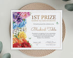 A printed certificate for 1st Prize Rainbow Tea Table Decoration styled on a woven round mat with eucalyptus leaves. The certificate features colorful watercolor flowers on the left side and elegant gold text on a white background.