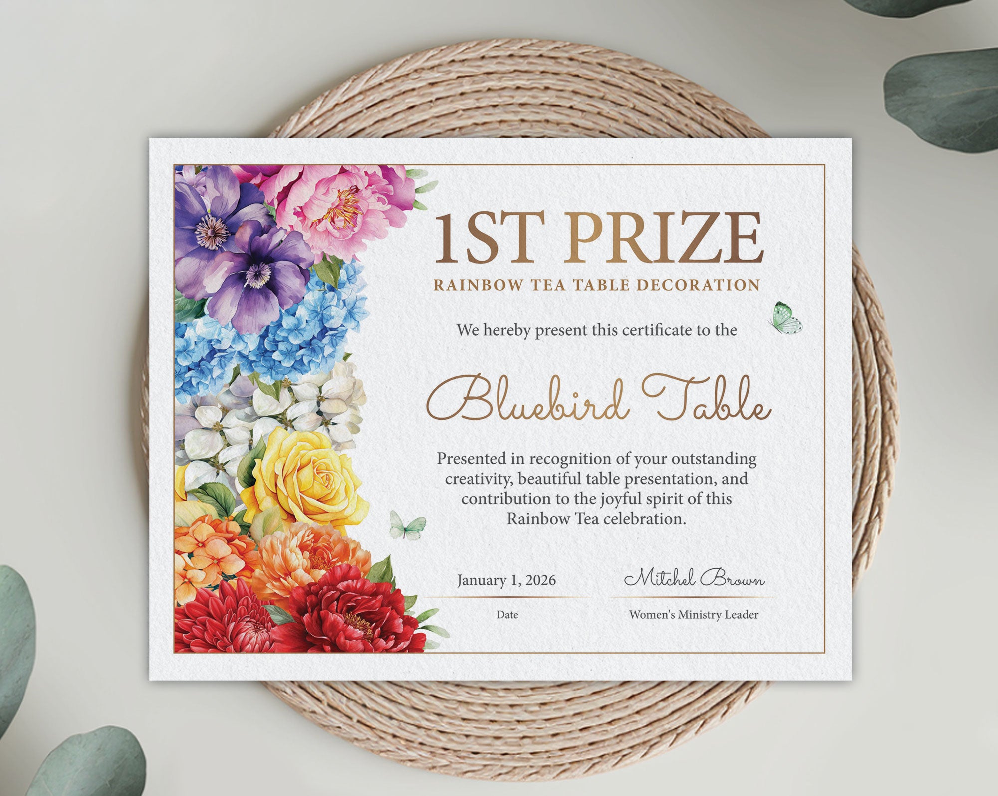 A printed certificate for 1st Prize Rainbow Tea Table Decoration styled on a woven round mat with eucalyptus leaves. The certificate features colorful watercolor flowers on the left side and elegant gold text on a white background.