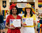 Two women standing in a decorated hall, each holding a prize certificate. The woman on the left wears a red lace dress and red fascinator, and the woman on the right wears a yellow hat and yellow blouse.