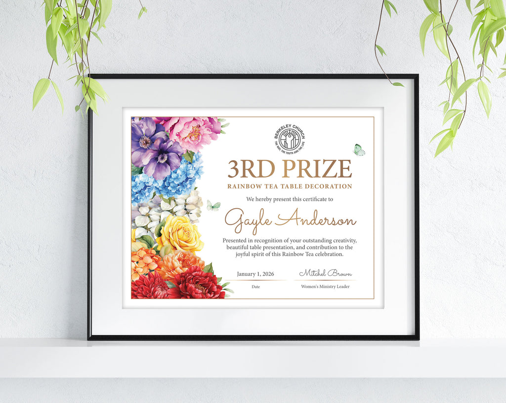A framed personalized certificate print leaning against a white wall, with green plant leaves hanging from above. The certificate has a colorful floral border and gold text reading '3rd Prize Rainbow Tea Table Decoration' and the name Gayle Anderson.