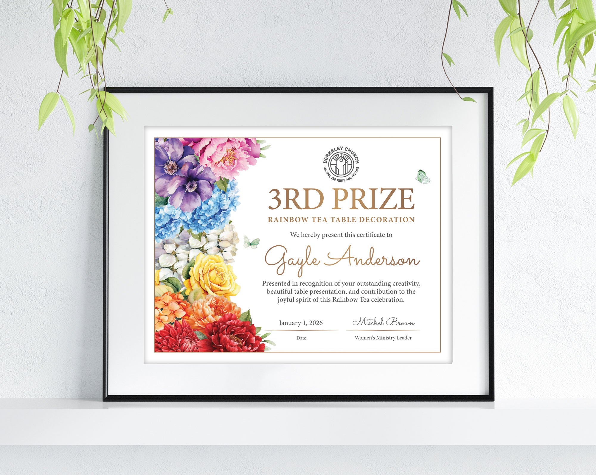 A framed personalized certificate print leaning against a white wall, with green plant leaves hanging from above. The certificate has a colorful floral border and gold text reading '3rd Prize Rainbow Tea Table Decoration' and the name Gayle Anderson.