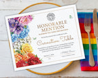A printed certificate card with floral artwork and gold text, styled on a gold-rimmed plate with a fork and knife on a rainbow napkin over a white wood surface.