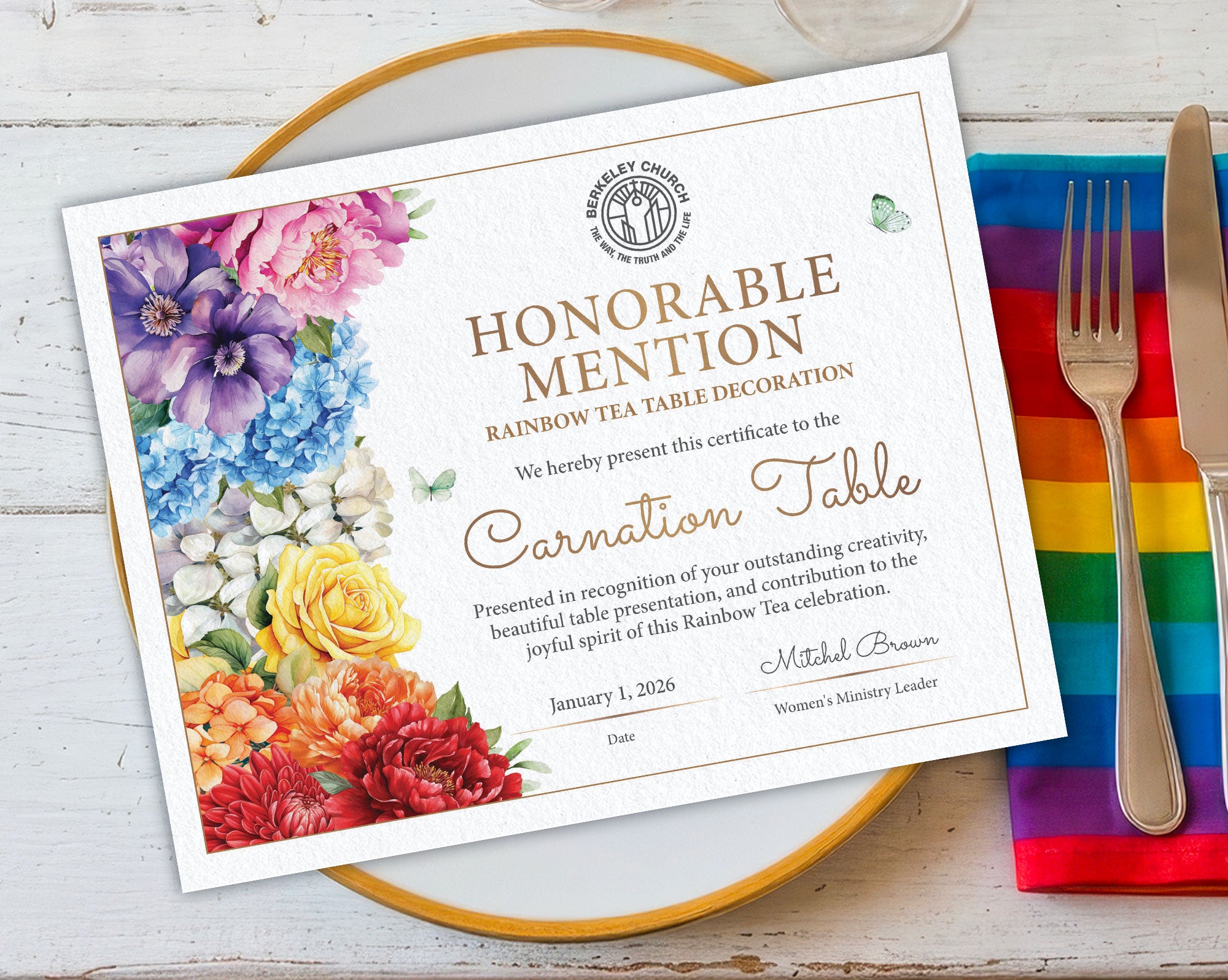 A printed certificate card with floral artwork and gold text, styled on a gold-rimmed plate with a fork and knife on a rainbow napkin over a white wood surface.