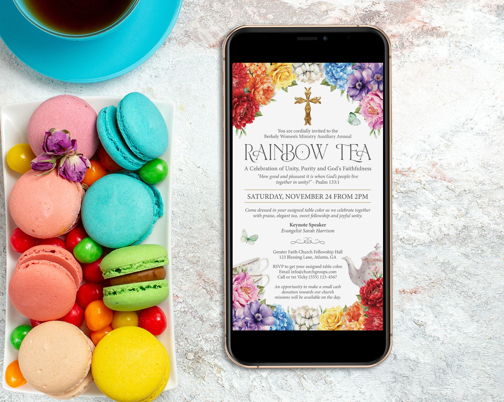 a black mobile phone showing a church rainbow tea invitation, laying on a marble surface next to a tray fill of colorful macarons and a blue tea cup