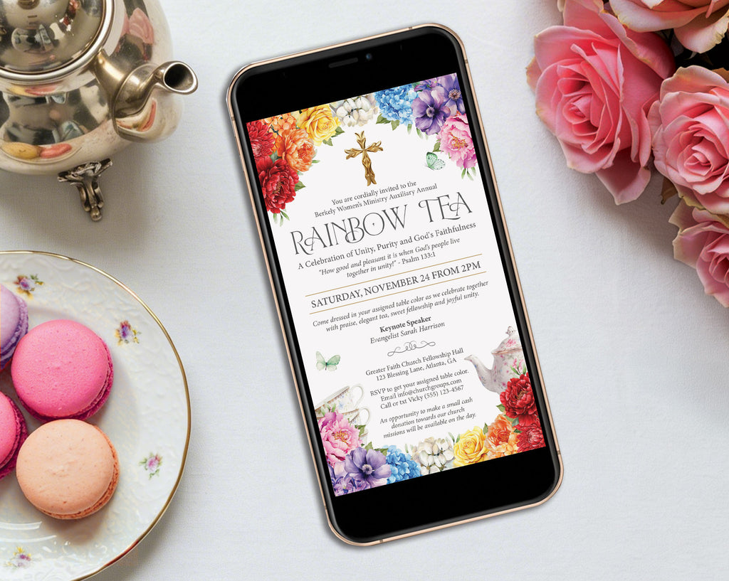 a bright rainbow tea mobile showing on the screen of a black mobile phone laying on a grey table with a silver teapot, some pink roses and macarons on a plate