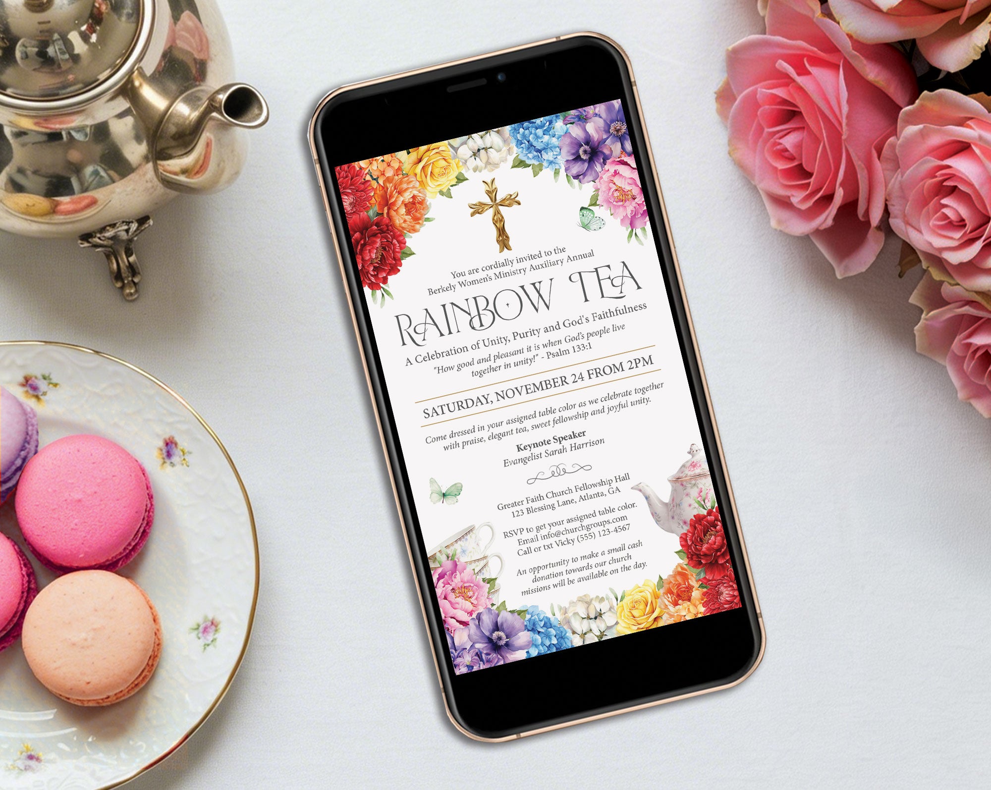 a bright rainbow tea mobile showing on the screen of a black mobile phone laying on a grey table with a silver teapot, some pink roses and macarons on a plate