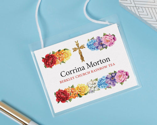 A rainbow tea name badge for a church event, with bright flowers and gold cross graphics, in a plastic holder with neck straps lying on a blue background next to a pen