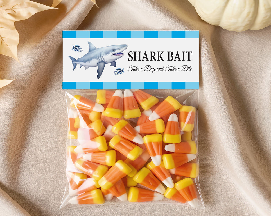 A clear plastic bag filled with candy corn treats, sealed with a printed label that reads "SHARK BAIT" and includes a shark illustration. The bag is photographed on draped beige fabric with decorative leaves and a small pumpkin in the background.