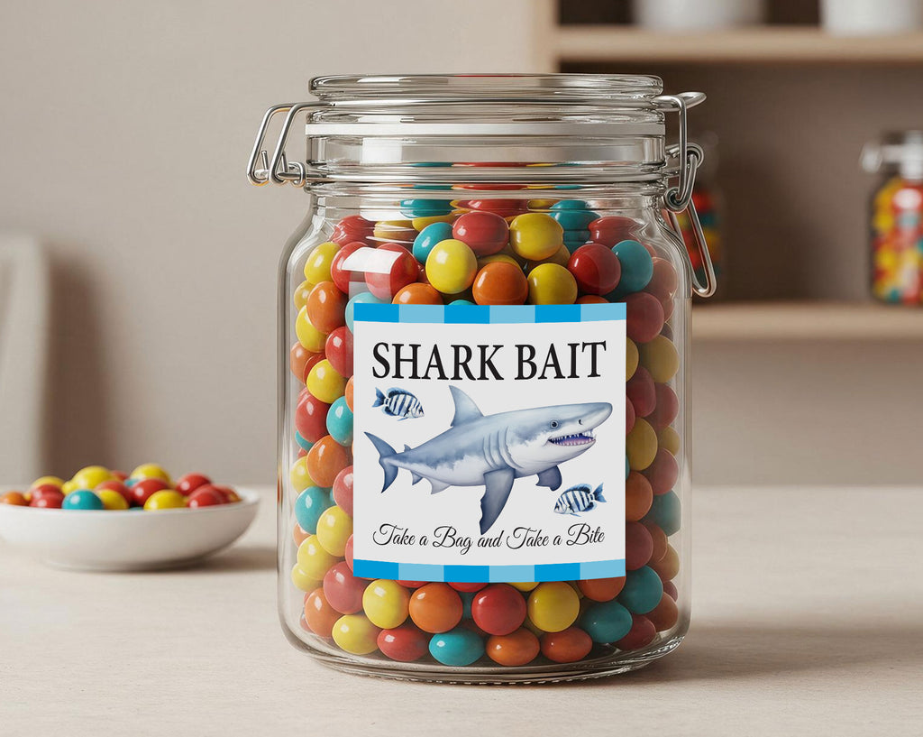 A clear glass jar filled with colorful round candies, labeled 'SHARK BAIT' with a shark illustration and the phrase 'Take a Bay and Take a Bite.' The jar sits on a light surface with a small bowl of similar candies blurred in the background.