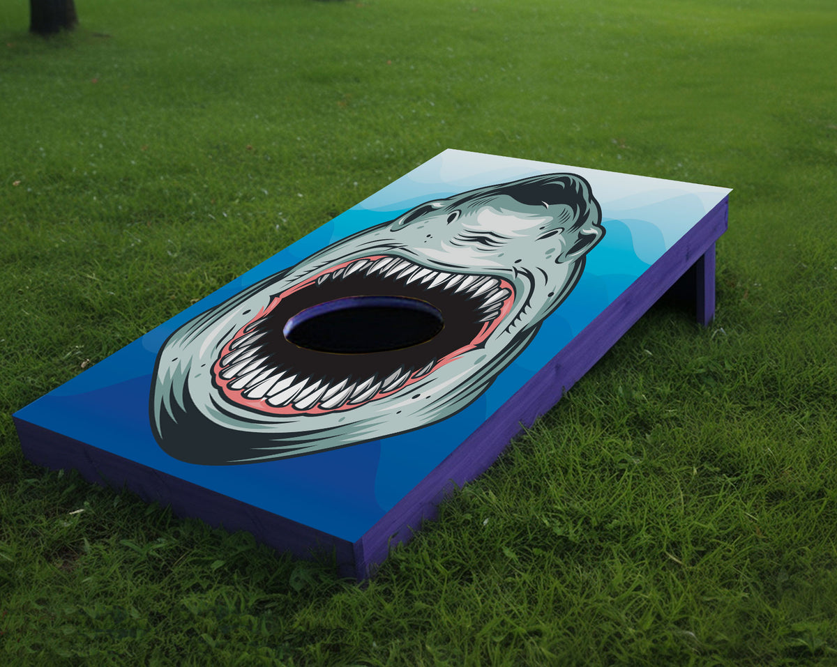 A cornhole board set outdoors on a green grass lawn, featuring a painted shark mouth graphic with an open mouth hole in the center.