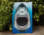 A rectangular canvas print featuring a stylized open shark face with sharp teeth, leaning against a stone surface outdoors in front of green flowering bushes. The artwork has a blue gradient background.