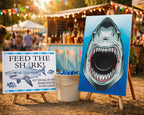 Outdoor party game setup featuring a large shark-themed canvas print on an easel and a sign that reads 'FEED THE SHARK!' with a bucket of blue balls in the center, set against a blurred festival background.