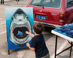 A child stands in a parking lot, reaching toward a blue square inside the mouth of a large illustrated shark on a freestanding sign. A red Honda station wagon is behind the child, and a table with blue items is to the right.