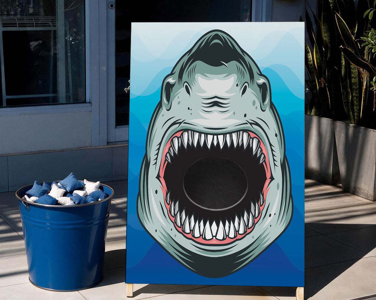 A vertical printed board featuring a large cartoon shark face with an open mouth, set outdoors beside a blue bucket filled with blue-and-white starfish. The scene includes a building entrance, tiled floor, and potted plants.