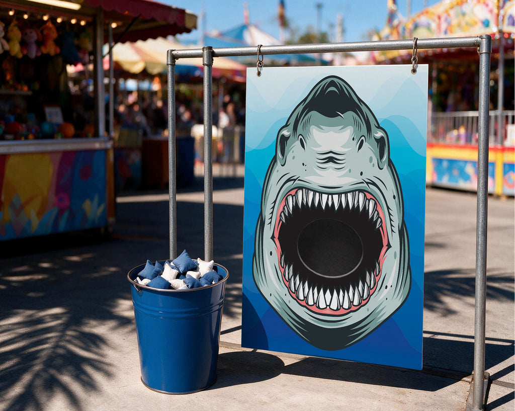 A carnival-style shark head target sign on a metal frame next to a blue bucket filled with blue and white targets, set outdoors at a fair with blurred rides and stalls in the background.