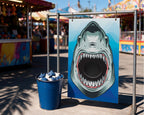A carnival-style shark head target sign on a metal frame next to a blue bucket filled with blue and white targets, set outdoors at a fair with blurred rides and stalls in the background.