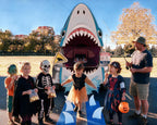 A shark-themed car decoration design with a gray car shown from the back, featuring a shark face in the trunk opening, white teeth, blue water splashes at the bottom, surrounded by kids in Halloween costumes and an adult in shorts and t-shirt holding a fishing rod