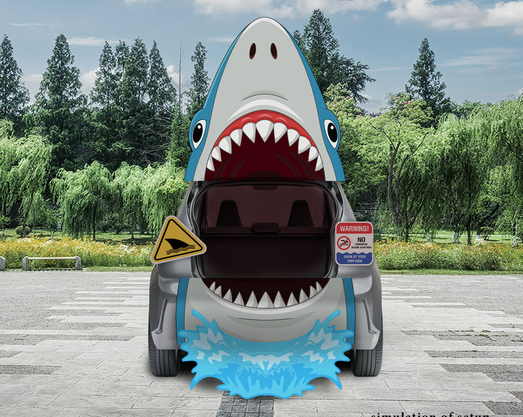 A car with a shark graphic on the front, showing an open trunk and wide-open mouth with white teeth, parked outdoors on a paved area with trees and sky in the background. 