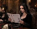 A woman in a dark gothic-style room holding a newspaper, wearing a black velvet dress with lace trim and layered necklaces, with candles and ornate furniture in the background.