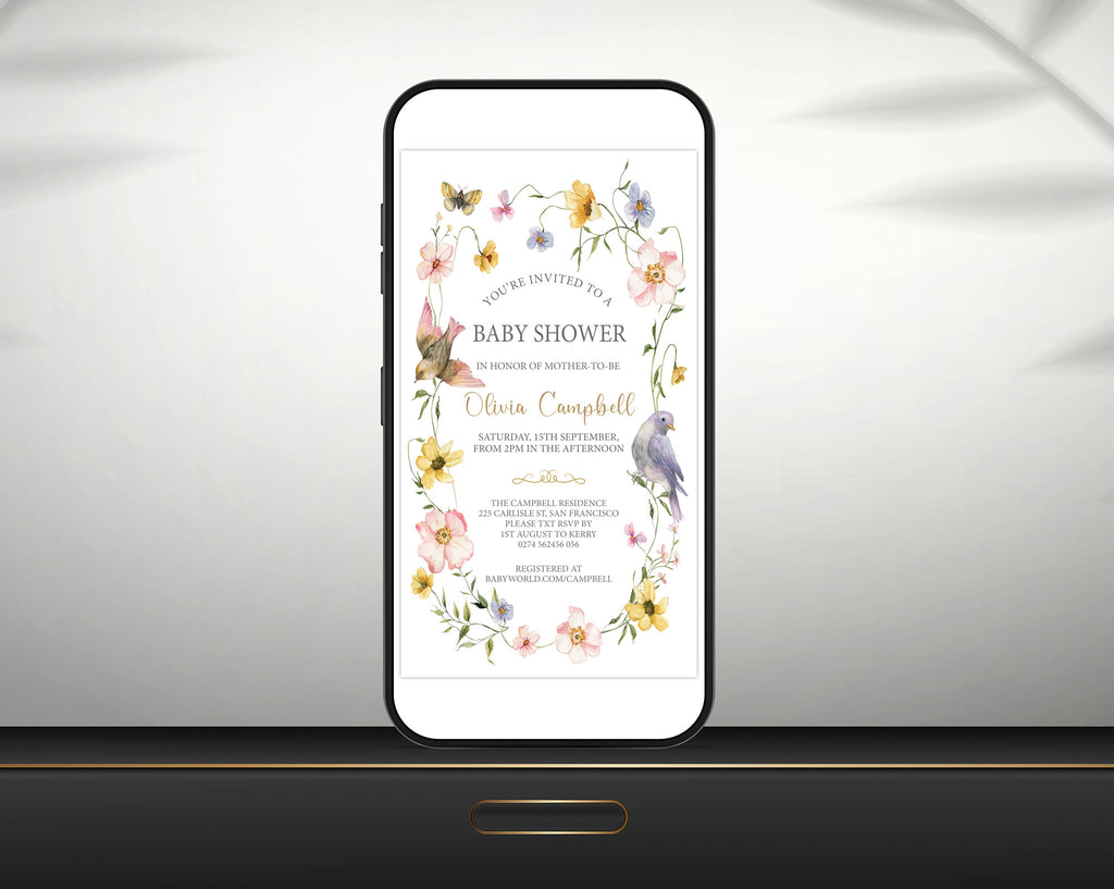 A mobile phone shows a baby shower invitation. The design includes watercolour flowers, butterflies, and birds, leaning up against a grey wall on a black table