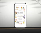 A mobile phone shows a baby shower invitation. The design includes watercolour flowers, butterflies, and birds, leaning up against a grey wall on a black table
