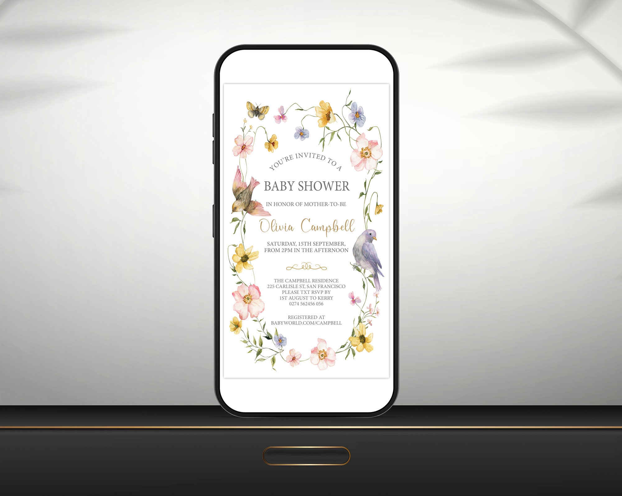 A mobile phone shows a baby shower invitation. The design includes watercolour flowers, butterflies, and birds, leaning up against a grey wall on a black table