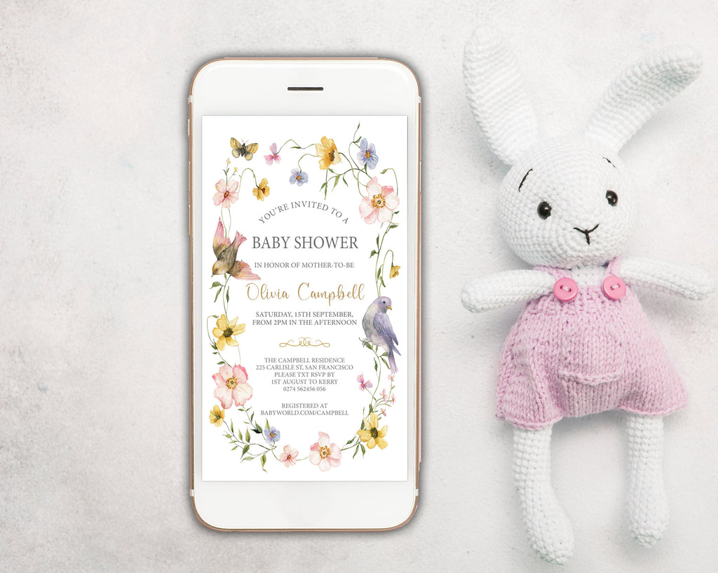a white mobile phone showing a wildflower and bird style baby shower invitation on the screen, with a white stuffed bunny in pink dungarees next to it