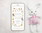 a white mobile phone showing a wildflower and bird style baby shower invitation on the screen, with a white stuffed bunny in pink dungarees next to it