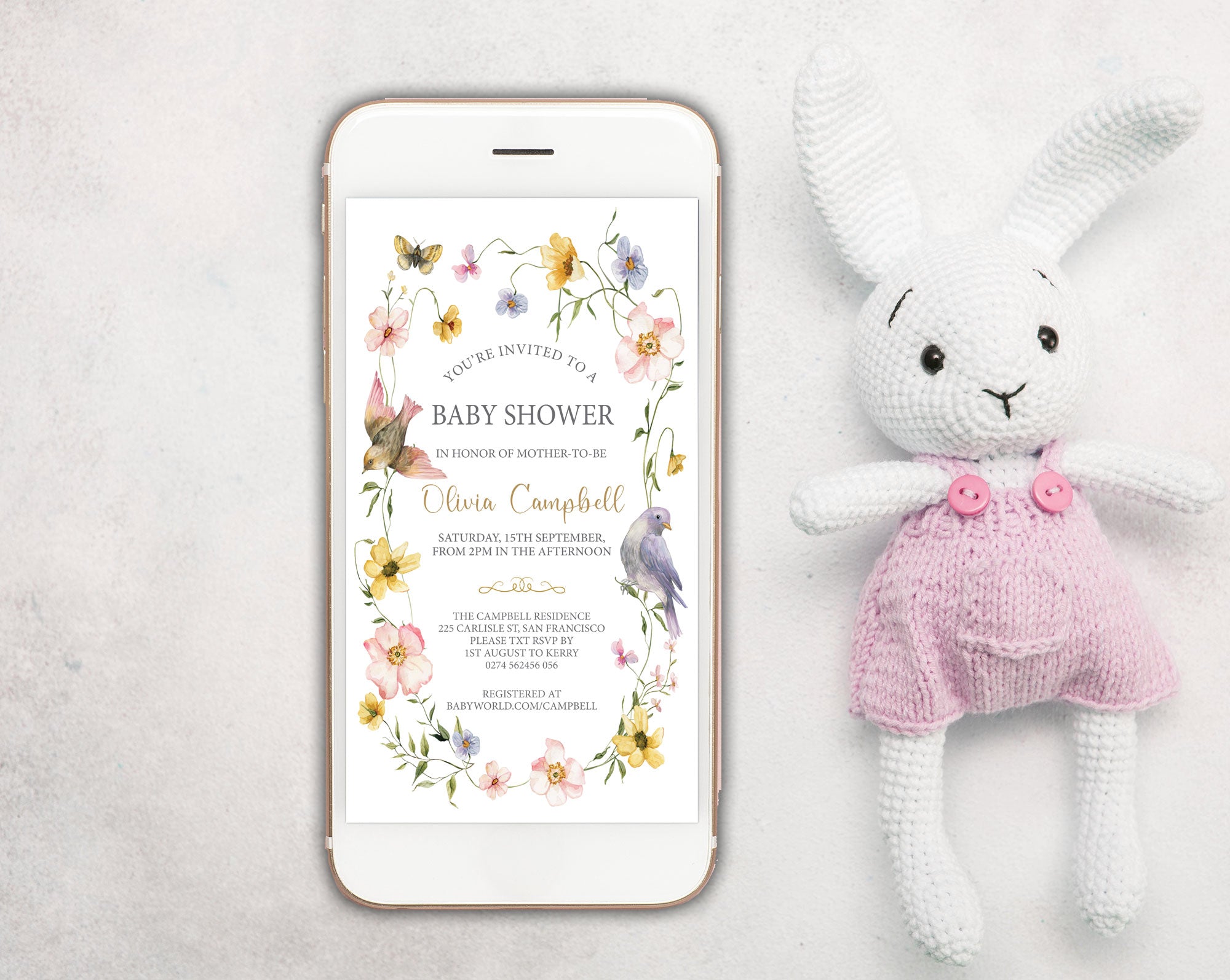 a white mobile phone showing a wildflower and bird style baby shower invitation on the screen, with a white stuffed bunny in pink dungarees next to it