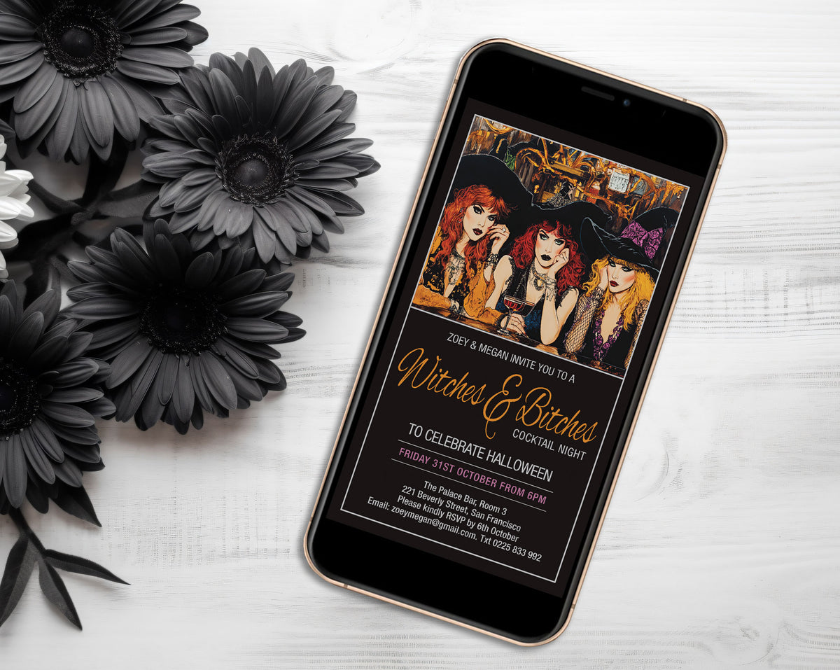 A smartphone lying on a white wood surface, displaying a Halloween invitation design with witch illustrations and gold script text. Black artificial flowers are arranged around it on the left.