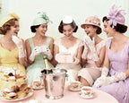 Five women in vintage dresses and hats sitting around a table with tea and pastries, smiling.