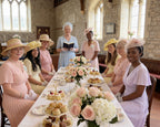 Group of women in formal attire sitting around a table with tea and pastries in a church setting.