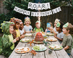 kids in dinosaur masks and fancy dress, sitting at a wooden table outside, with party sandwiches and a volcano cake with happy birthday bunting in the background