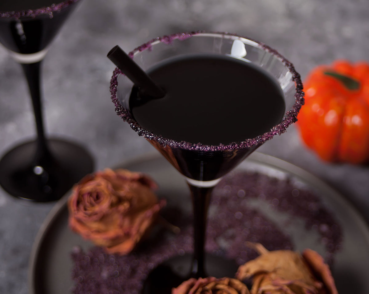 A Halloween-themed cocktail in a martini glass with a dark purple sugar-rimmed edge, filled with black liquid and a black straw, set on a plate with dried roses and a small orange pumpkin against a gray background.