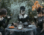 Three women in gothic black lace dresses sit around a table outdoors, holding teacups in a moody, Halloween-themed lifestyle setting with skulls and pumpkins on the tablecloth.
