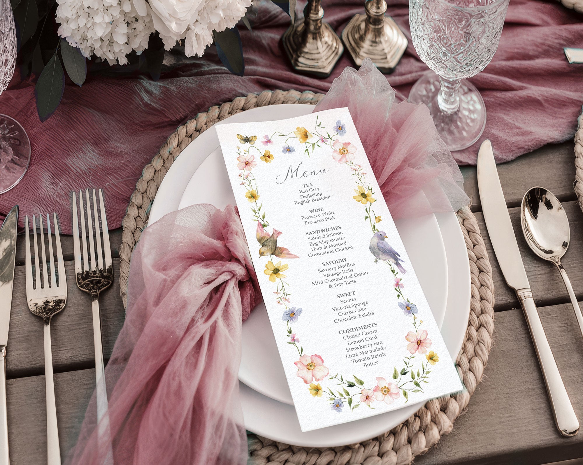 pretty baby shower menu with birds and wildflowers laying on a white plate on a pink gauzy cloth, on a wooden table set with cutlery